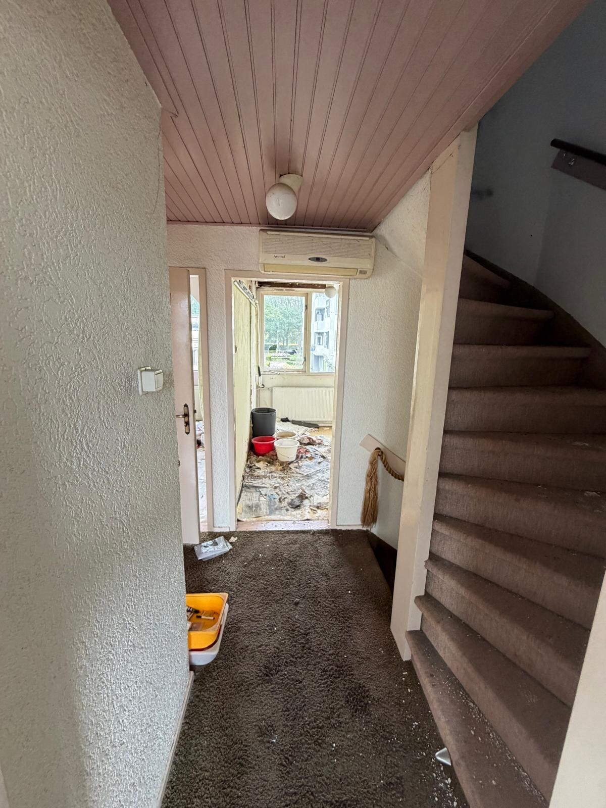 Overloop met roze schrootjesplafond, bruin tapijt op de trap en puin op de vloer, voor het strippen