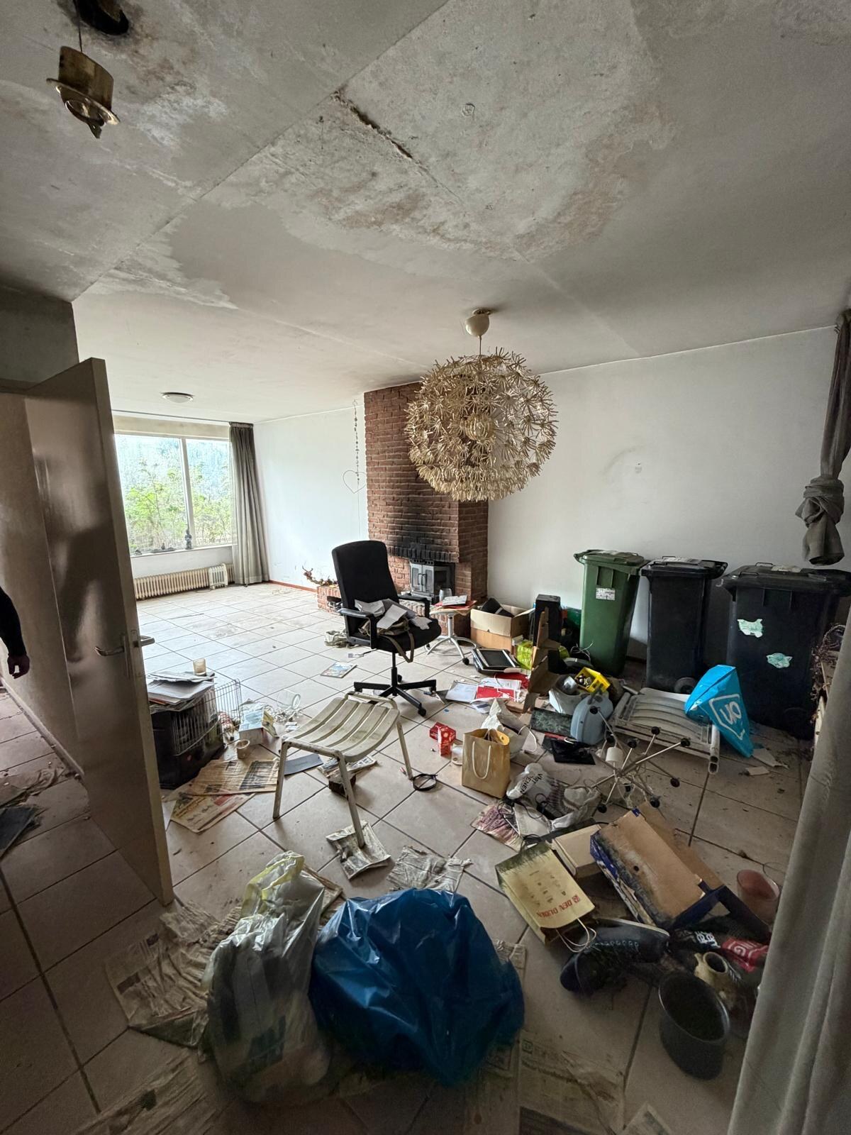 Woonkamer vol spullen en puin, met een gemetselde schouw en een kroonluchter, voor het strippen